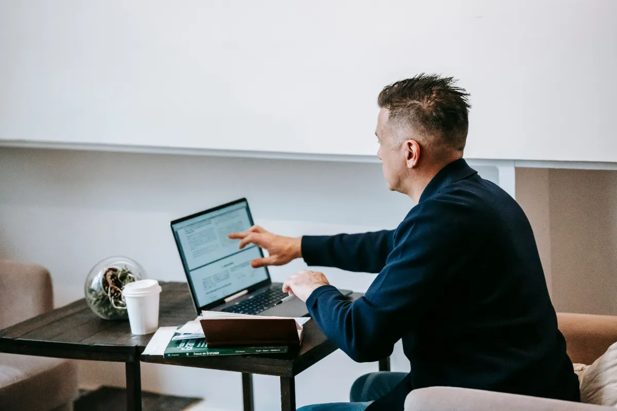A man at a low table pointing at a text-heavy page on his laptop screen